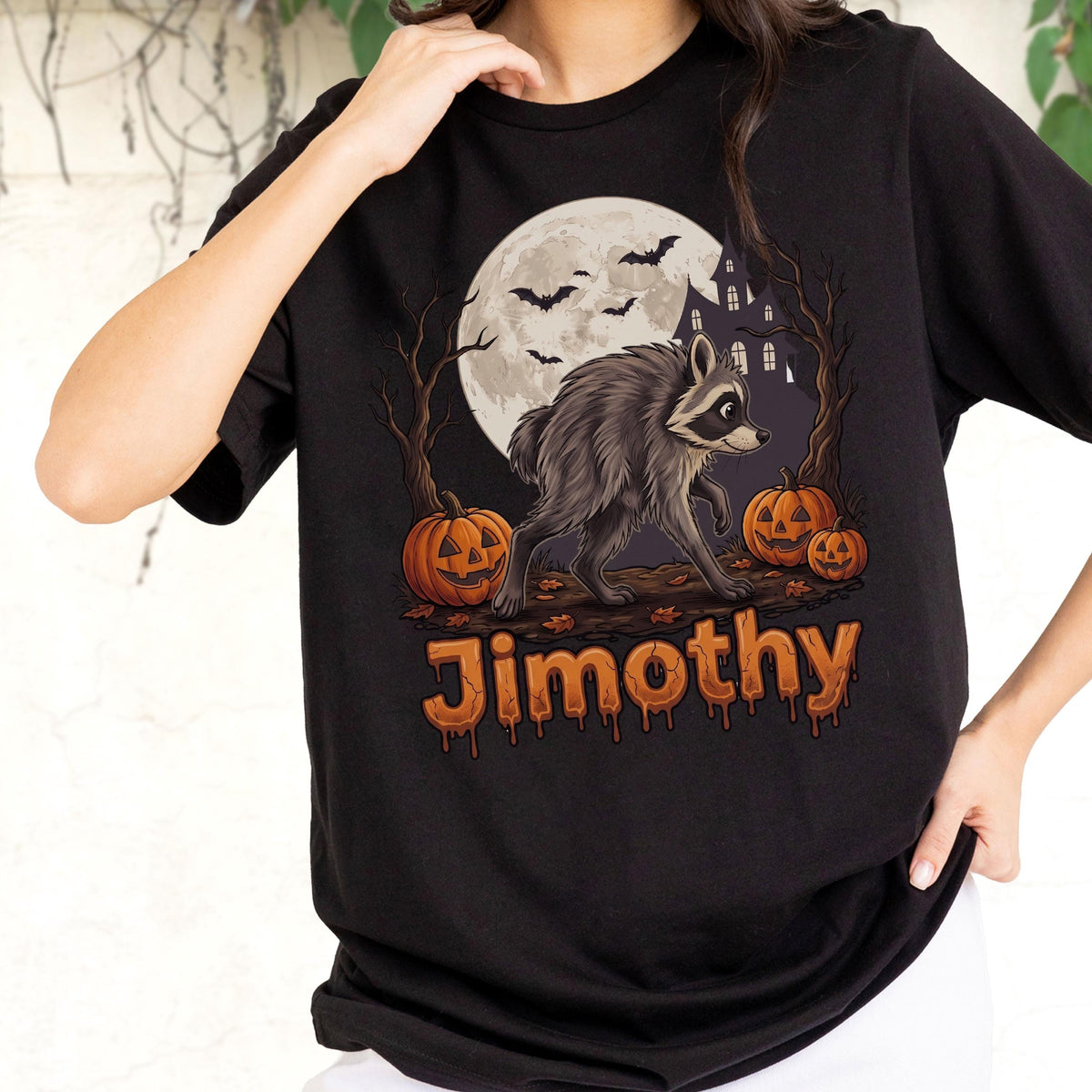 May include: A black t-shirt featuring a graphic of a raccoon standing in front of a full moon, bare trees, and a haunted house. Three orange jack-o'-lanterns are on the ground with bats flying above. The name Jimothy is printed in orange.