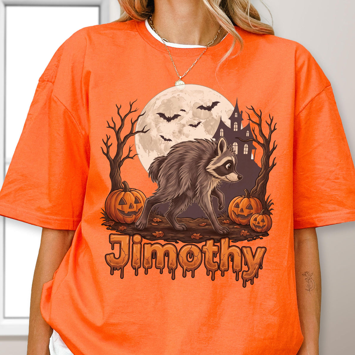 May include: An orange t-shirt featuring a graphic of a raccoon walking in front of a full moon, a haunted house, bare trees, and three jack-o'-lanterns. Bats fly in the sky, and the name Jimothy is printed in a dripping, orange-brown font below.
