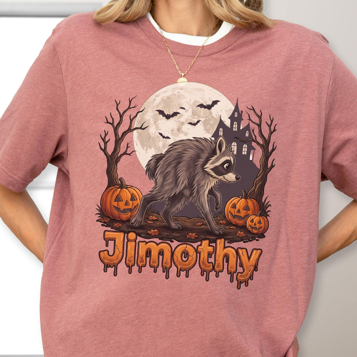 May include: A dusty rose t-shirt featuring a graphic of a raccoon walking in front of a full moon, a haunted house, bare trees, and three jack-o'-lanterns. The text "Jimothy" is printed in a dripping orange font at the bottom of the design.