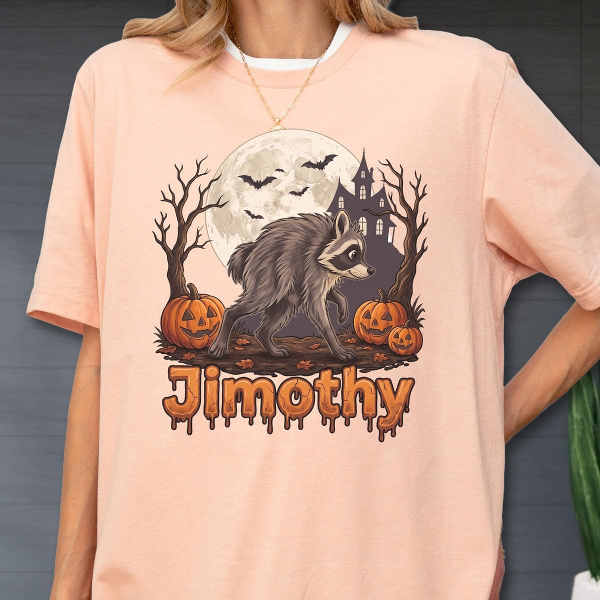 May include: A peach-colored t-shirt featuring a graphic of a raccoon walking in front of a full moon, a haunted house, bare trees, and three jack-o'-lanterns. The word "Jimothy" is printed below the graphic in a dripping, orange-toned font.
