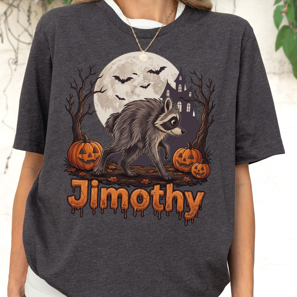 May include: A dark heather gray t-shirt featuring a graphic of a raccoon walking in front of a full moon, a haunted house, bare trees, and three jack-o'-lanterns. The name Jimothy is printed in orange, dripping, stylized lettering below the graphic.