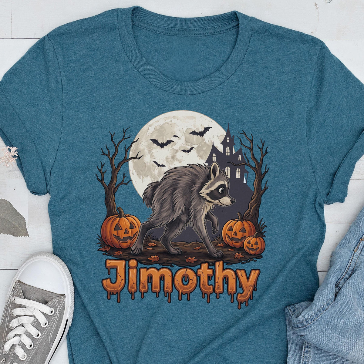 May include: A teal t-shirt featuring a graphic of a raccoon standing between two bare trees, three orange jack-o'-lanterns, a large full moon, bats, and a dark house silhouette. The name Jimothy is printed below in an orange, dripping font.
