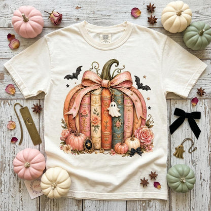 May include: A cream-colored short-sleeve t-shirt featuring a graphic of a pumpkin shaped like a stack of vintage books tied with a pink ribbon. The design includes a small ghost charm, bats, stars, and floral accents in shades of pink and blue.