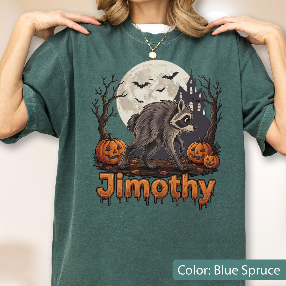 May include: A blue spruce t-shirt featuring a graphic of a raccoon, a full moon, bats, a haunted house, and jack-o'-lanterns. The text "Jimothy" is printed in orange below the graphic. The shirt has a crew neckline and short sleeves.