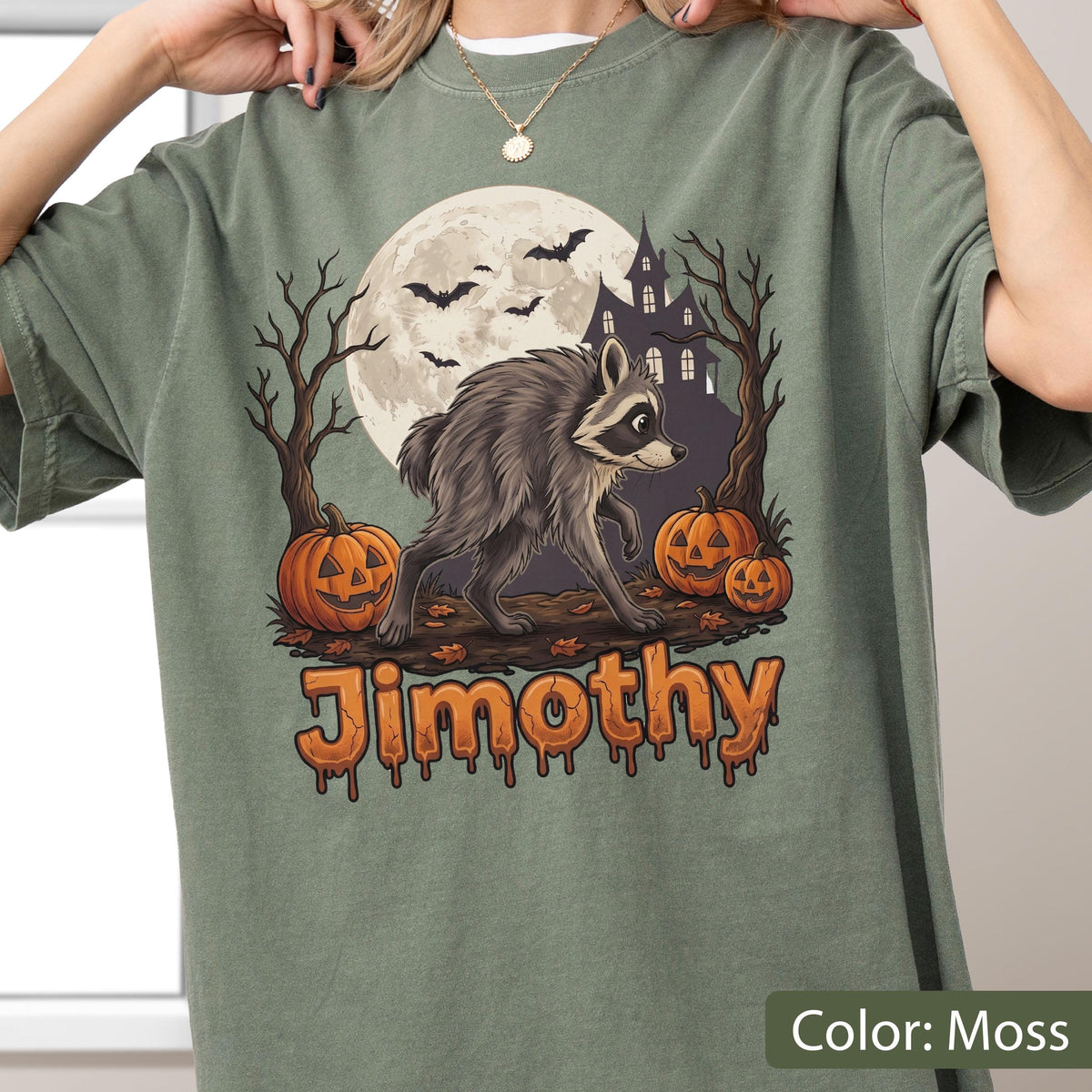 May include: A moss green t-shirt featuring a graphic of a raccoon walking in front of a full moon, a haunted house, bare trees, and three jack-o'-lanterns. The name Jimothy is printed in orange, dripping-style lettering at the bottom of the design.