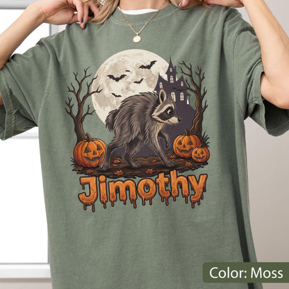 May include: A moss green t-shirt featuring a graphic of a raccoon walking in front of a full moon, a haunted house, bare trees, and three jack-o'-lanterns. The name Jimothy is printed in orange, dripping-style lettering at the bottom of the design.