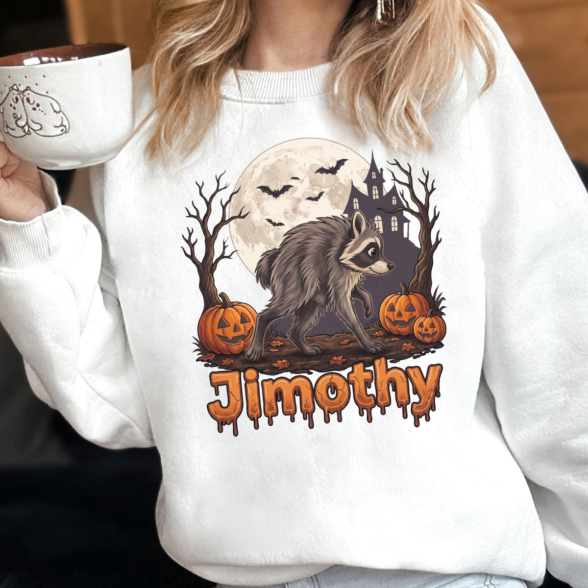 May include: A white sweatshirt featuring a graphic of a raccoon walking in front of a full moon, a haunted house, bare trees, and jack-o'-lanterns. The name "Jimothy" is printed below in orange, dripping-style lettering. The design has a spooky theme.