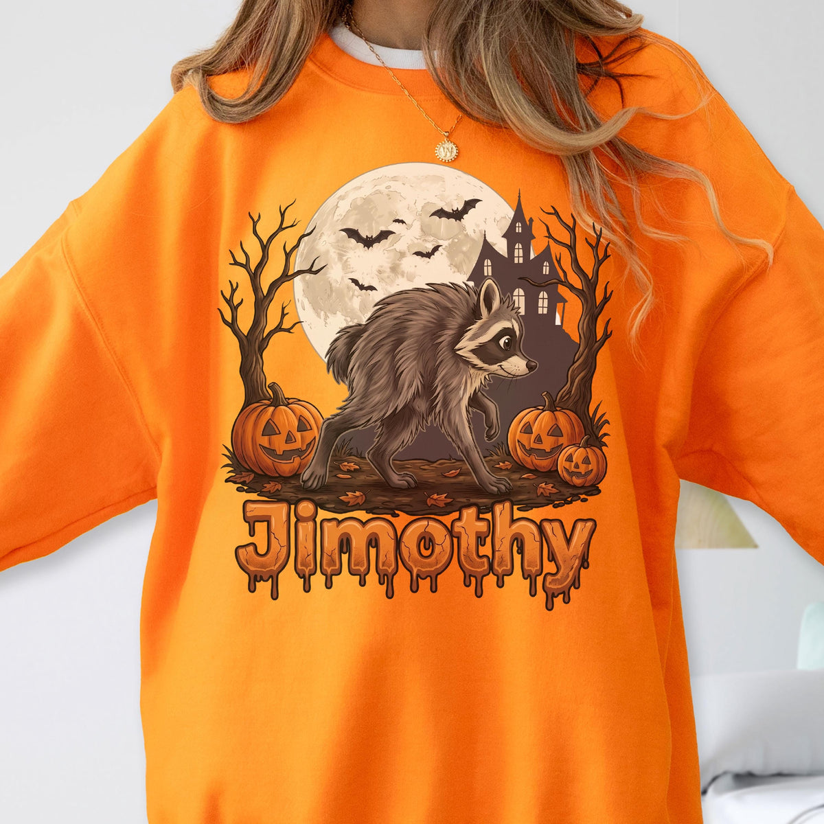 May include: An orange sweatshirt featuring a graphic of a raccoon walking in front of a full moon, a haunted house, bare trees, and three jack-o'-lanterns. The name "Jimothy" is printed in a dripping, orange, Halloween-style font below the graphic.