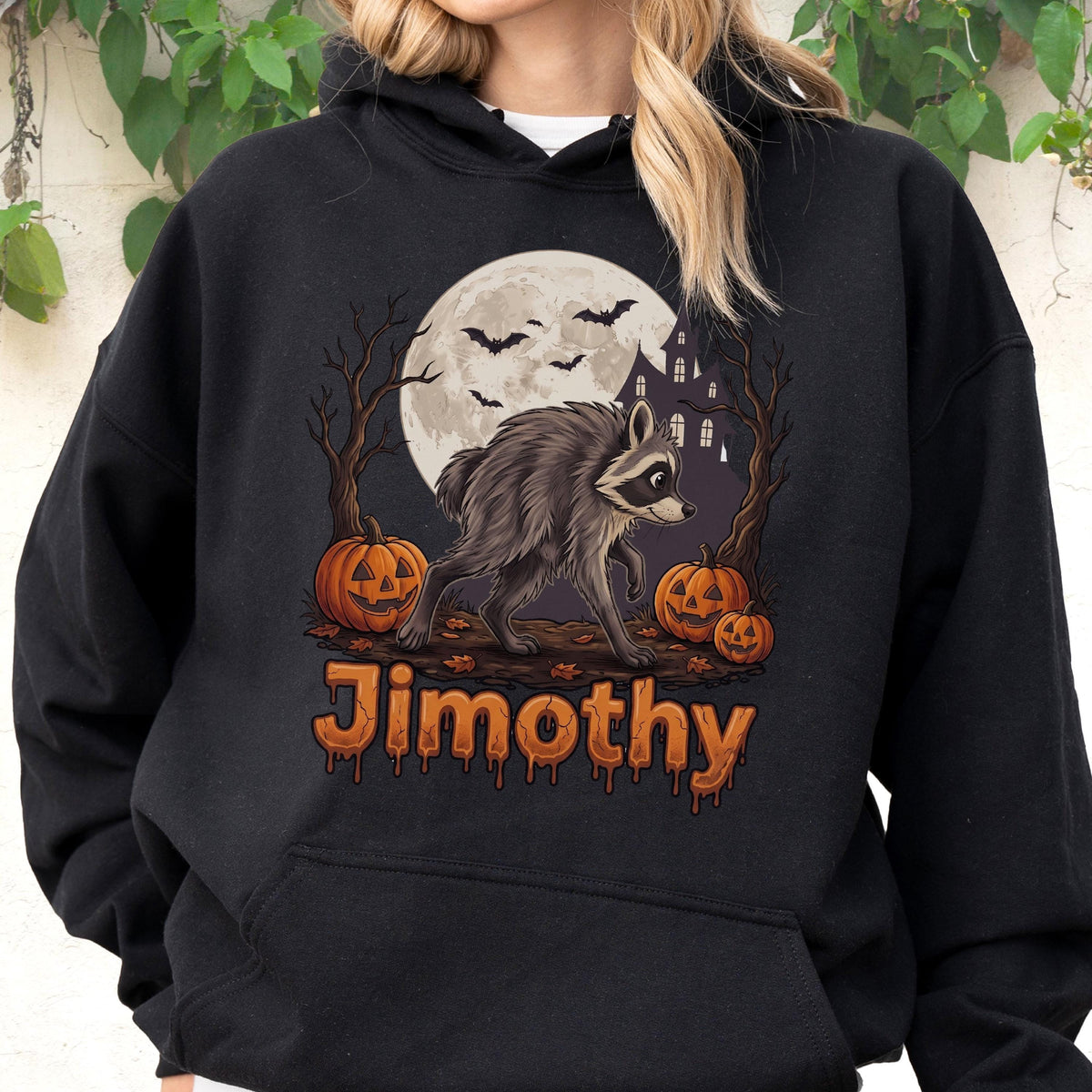 May include: A black hooded sweatshirt featuring a graphic print of a raccoon standing in front of a full moon, a haunted house, bare trees, and three jack-o'-lanterns. The name Jimothy is printed below the graphic in an orange, dripping font.