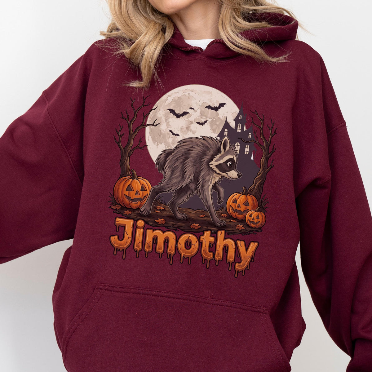 May include: A maroon hooded sweatshirt featuring a graphic print of a raccoon standing on a log, surrounded by three jack-o'-lanterns, bare trees, bats, and a full moon. The name Jimothy is printed in orange, dripping-style lettering below.