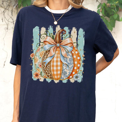 May include: A navy blue short-sleeve t-shirt featuring a central graphic of a pumpkin decorated with various patterns including leopard print, orange gingham, and blue floral designs, topped with a large light blue bow and surrounded by flowers.