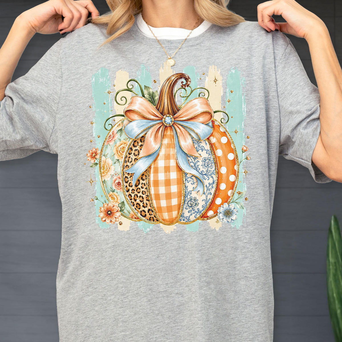 May include: A heather gray t-shirt featuring a central graphic of a pumpkin decorated with various patterns including leopard print, orange gingham, blue floral, and white polka dots, topped with a large light blue bow and surrounded by flowers.