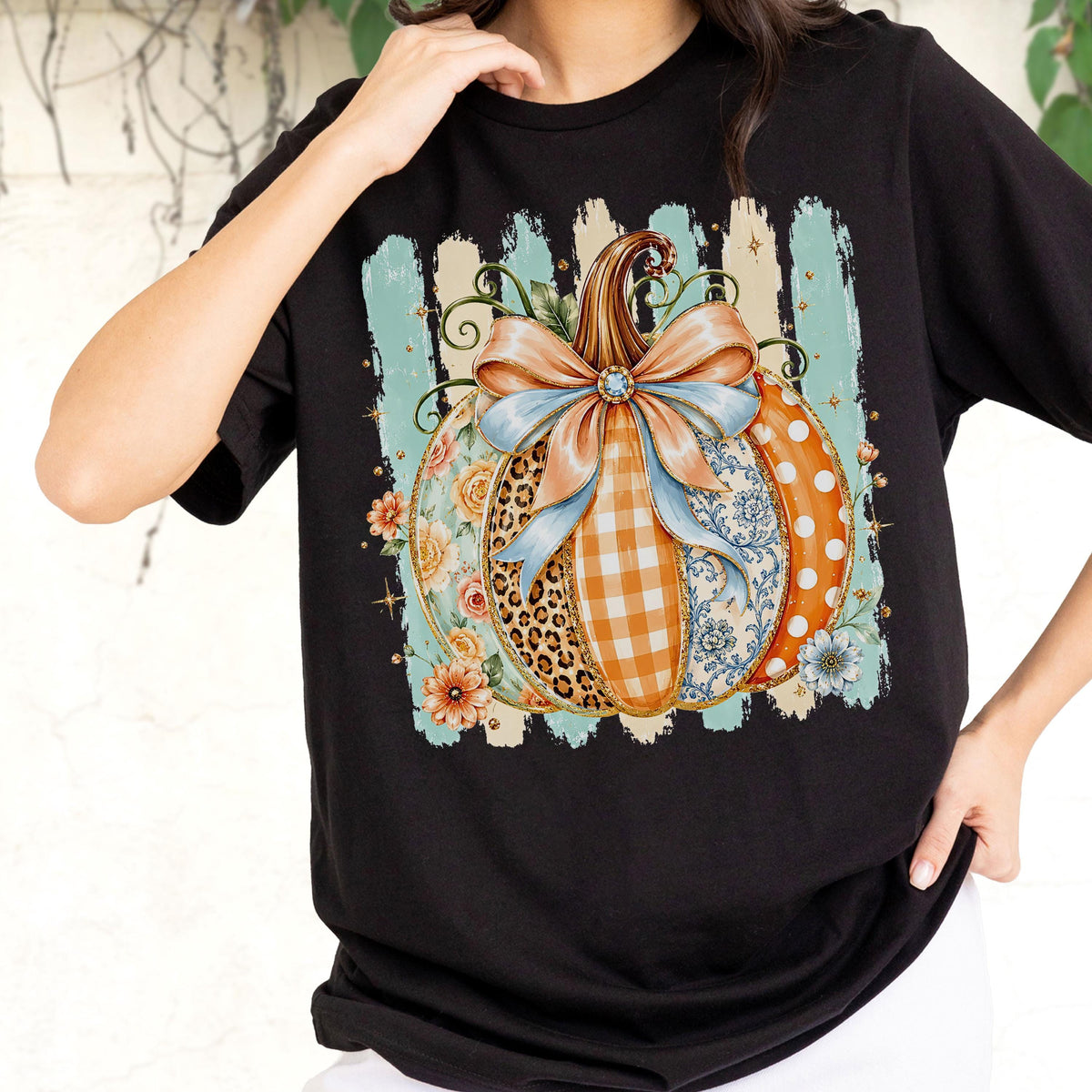 May include: A black t-shirt featuring a large graphic of a pumpkin decorated with various patterns including leopard print, orange gingham, and blue floral designs. The pumpkin is topped with a large light blue and peach bow and surrounded by flowers.