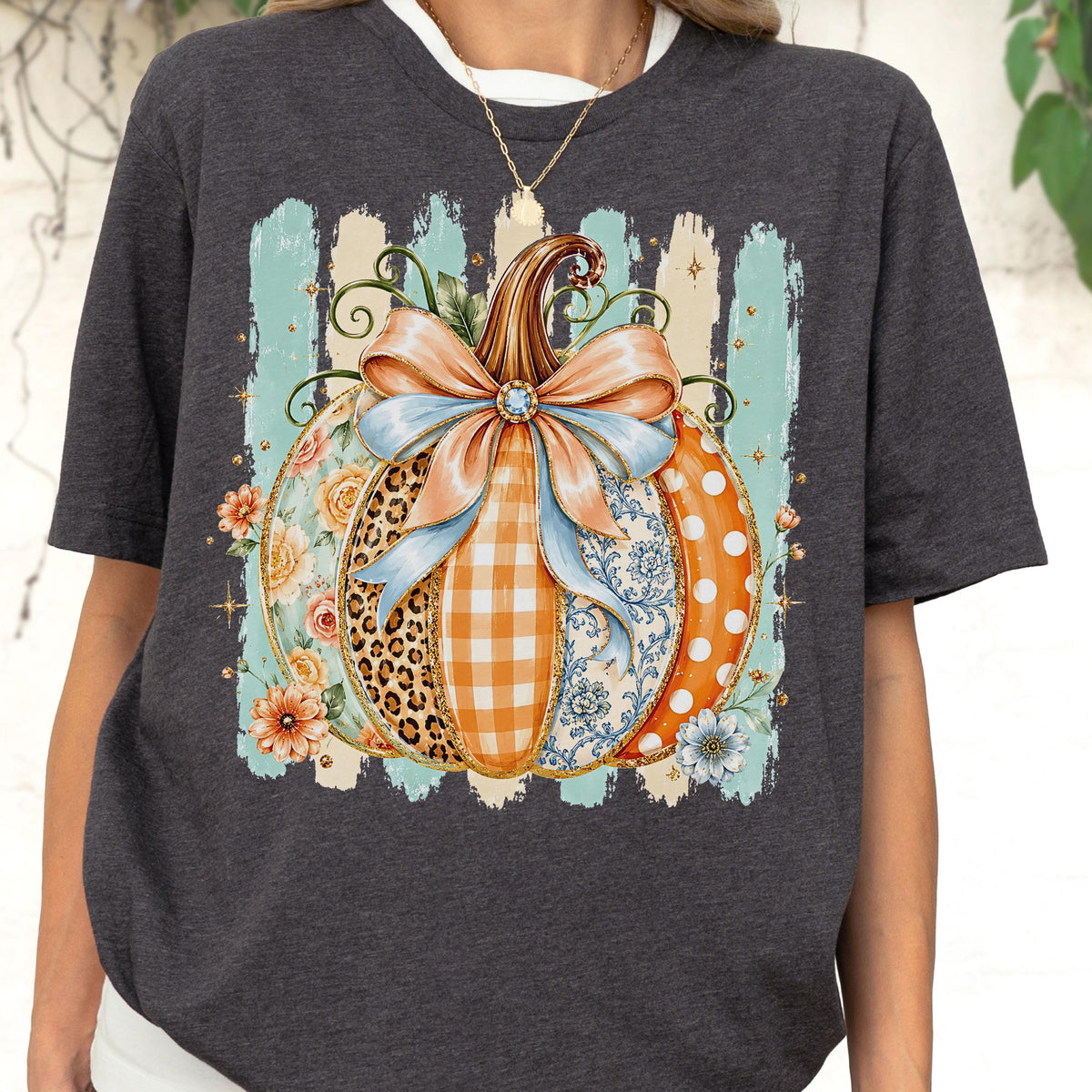 May include: A dark charcoal gray t-shirt featuring a large graphic of a pumpkin decorated with various patterns including leopard print, orange gingham, blue floral, and white polka dots, topped with a large light blue and peach bow.