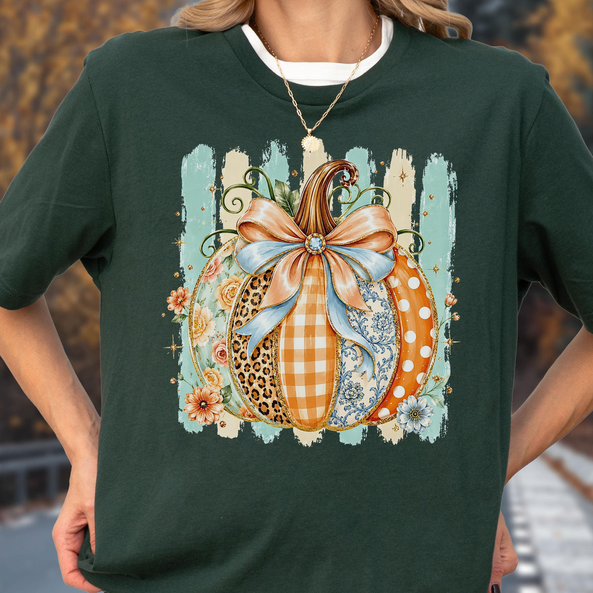 May include: A dark green t-shirt featuring a central graphic of a pumpkin decorated with various patterns including leopard print, orange gingham, blue floral, and white polka dots, topped with a large light blue bow and surrounded by flowers.