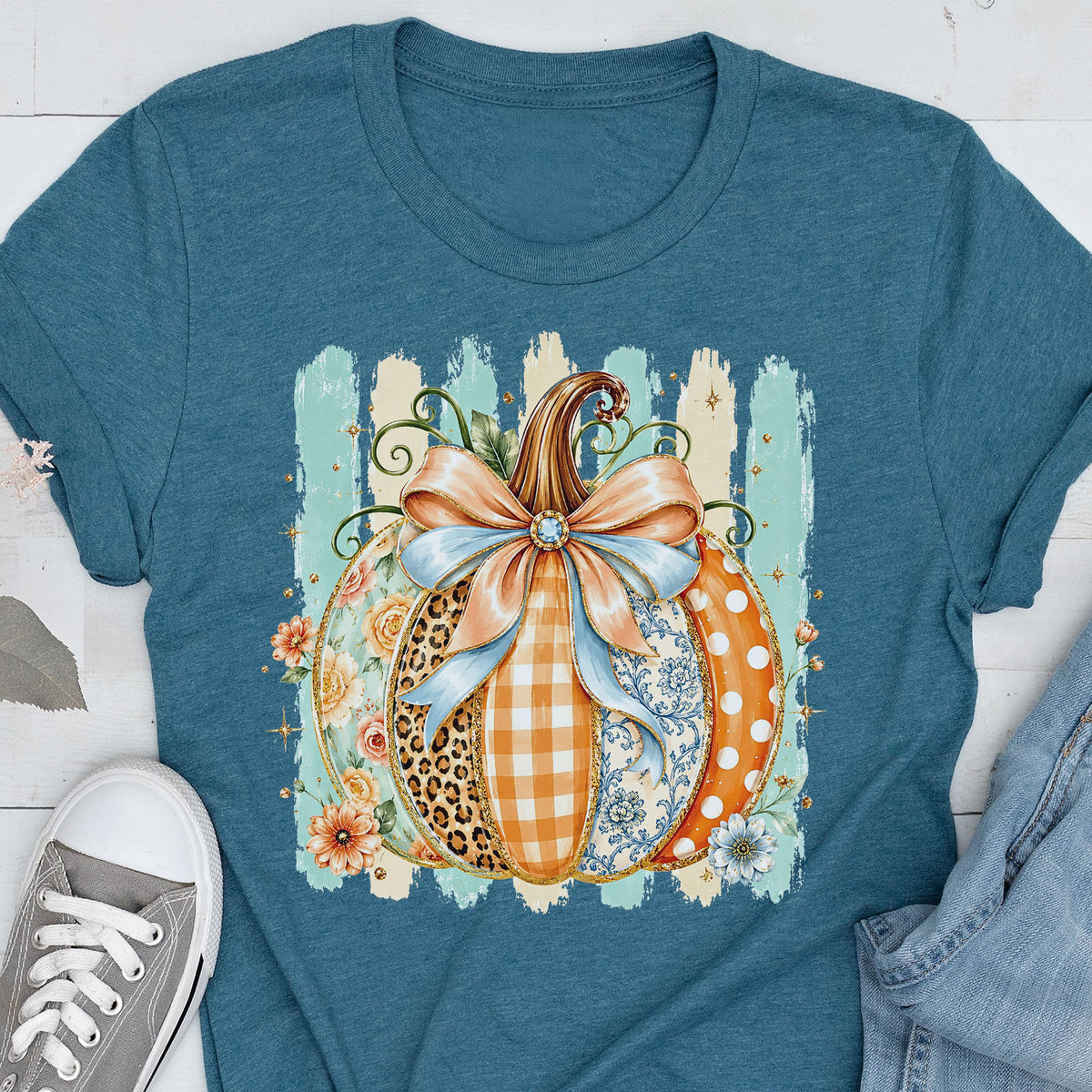 May include: A teal short-sleeve t-shirt featuring a central graphic of a pumpkin decorated with various patterns including leopard print, orange gingham, blue floral, and white polka dots, topped with a large light orange bow and flowers.