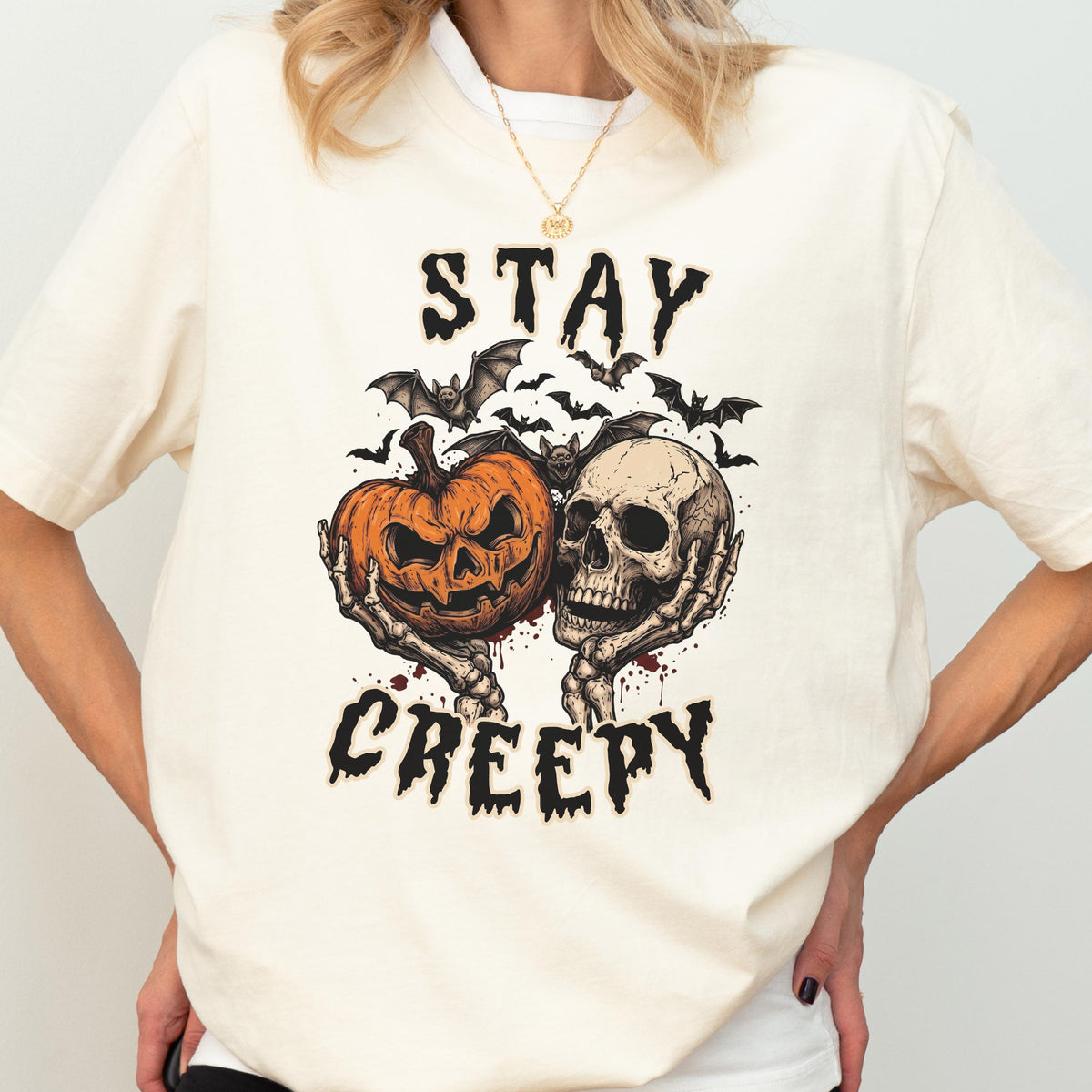May include: A cream-colored t-shirt featuring a graphic print of two skeletal hands holding a jack-o'-lantern and a human skull. Above and below the central illustration, the text reads STAY and CREEPY in a black, distressed, jagged font.