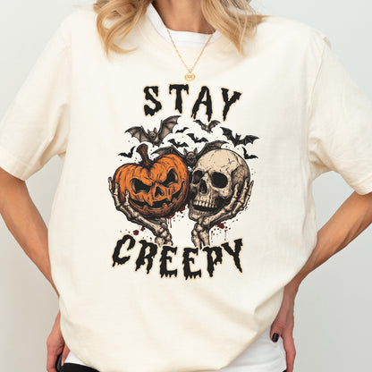 May include: A cream-colored t-shirt featuring a graphic print of two skeletal hands holding a jack-o'-lantern and a human skull. Above and below the central illustration, the text reads STAY and CREEPY in a black, distressed, jagged font.