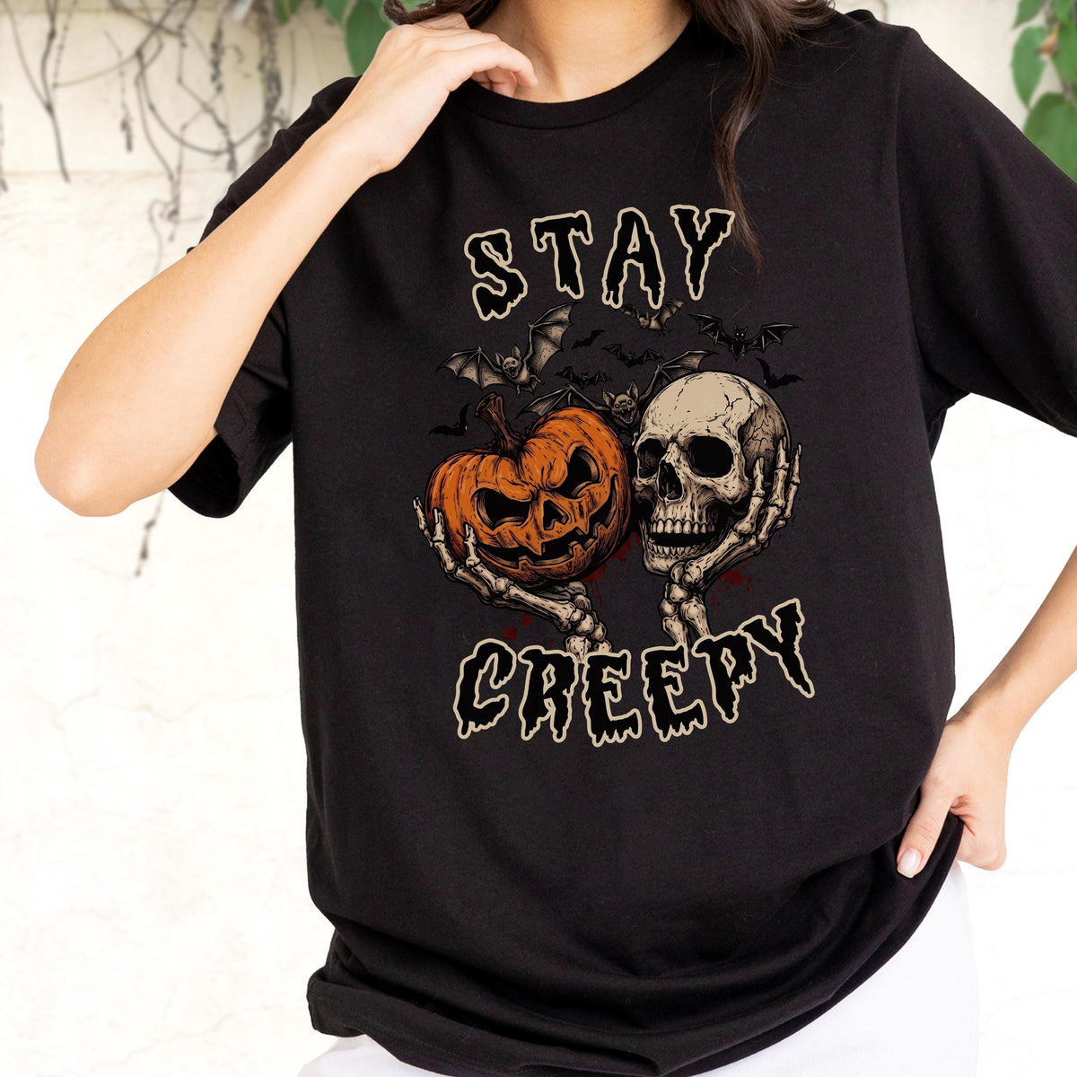 May include: A black short-sleeved t-shirt featuring a graphic print of a skeleton holding an orange jack-o'-lantern and a human skull. Above and below the illustration, the text reads STAY CREEPY in a stylized, dripping white font.
