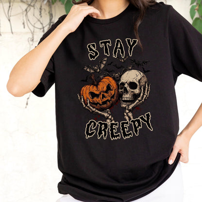 May include: A black short-sleeved t-shirt featuring a graphic print of a skeleton holding an orange jack-o'-lantern and a human skull. Above and below the illustration, the text reads STAY CREEPY in a stylized, dripping white font.