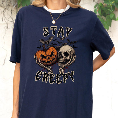 May include: A navy blue t-shirt featuring a graphic print of two skeletal hands holding a carved orange jack-o'-lantern and a human skull. Above and below the central illustration, the text reads STAY CREEPY in a jagged, dark font.