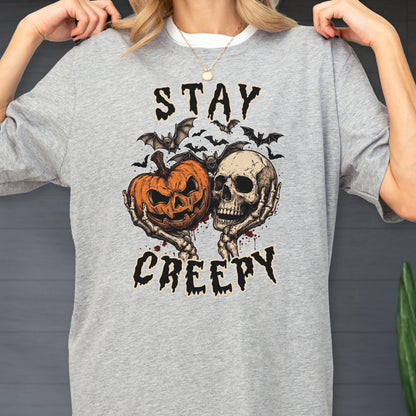 May include: A heather gray t-shirt featuring a graphic print of two skeletal hands holding an orange jack-o'-lantern and a human skull. Above and below the illustration, the text reads STAY CREEPY in a black, jagged, distressed font.