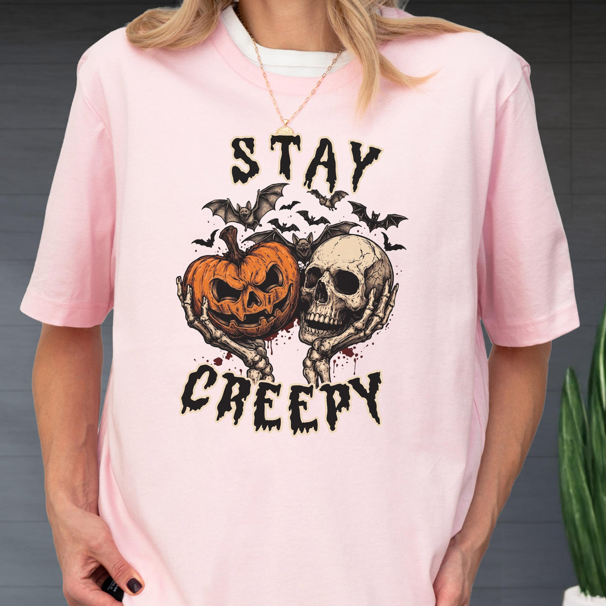 May include: A light pink t-shirt featuring a graphic print of a jack-o'-lantern and a human skull held by skeletal hands. Above and below the central illustration, the text reads STAY CREEPY in a jagged black font, surrounded by small bats.