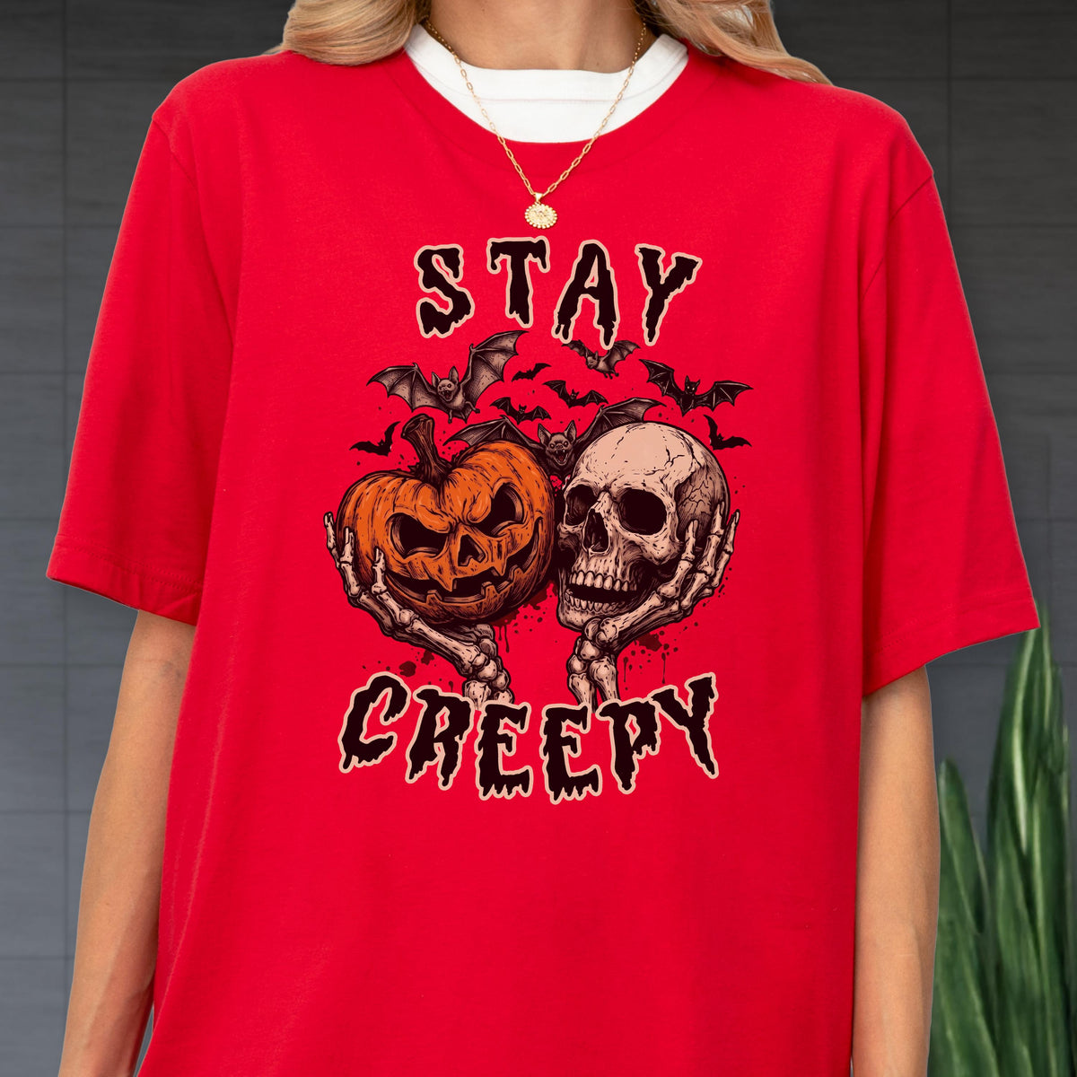 May include: A bright red short-sleeved t-shirt featuring a graphic print of two skeletal hands holding a jack-o'-lantern and a human skull, surrounded by bats and the text STAY CREEPY in a distressed black font, worn with a gold chain necklace.