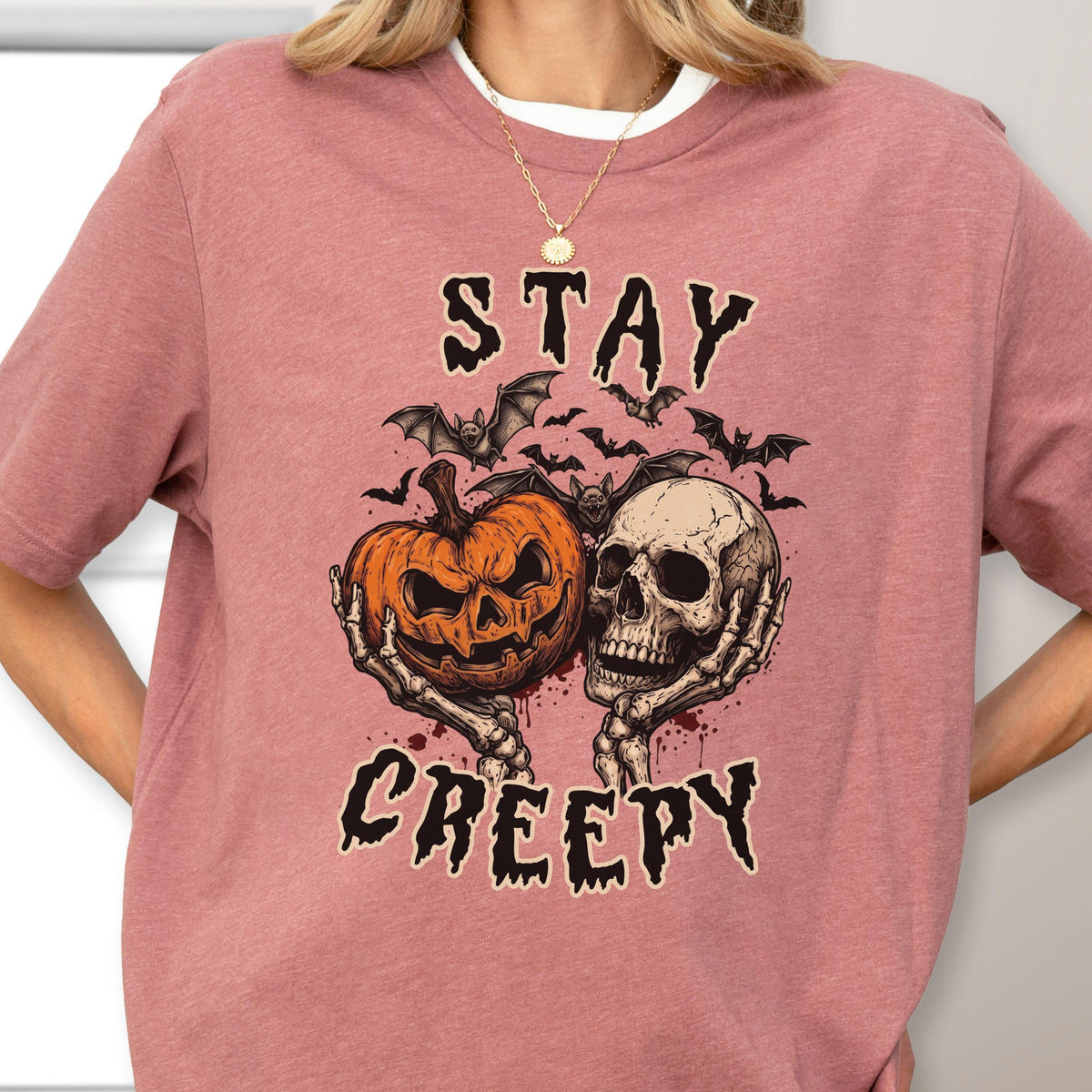 May include: A dusty rose t-shirt featuring a graphic print of two skeletal hands holding a carved orange jack-o'-lantern and a human skull. Above and below the central illustration, the text reads STAY and CREEPY in a jagged, dark font, surrounded by several small black bats.