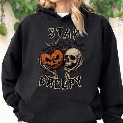 May include: A black hooded sweatshirt featuring a graphic print of two skeletal hands holding an orange jack-o'-lantern and a human skull. Above and below the central illustration, the text reads STAY and CREEPY in a stylized, dripping font.