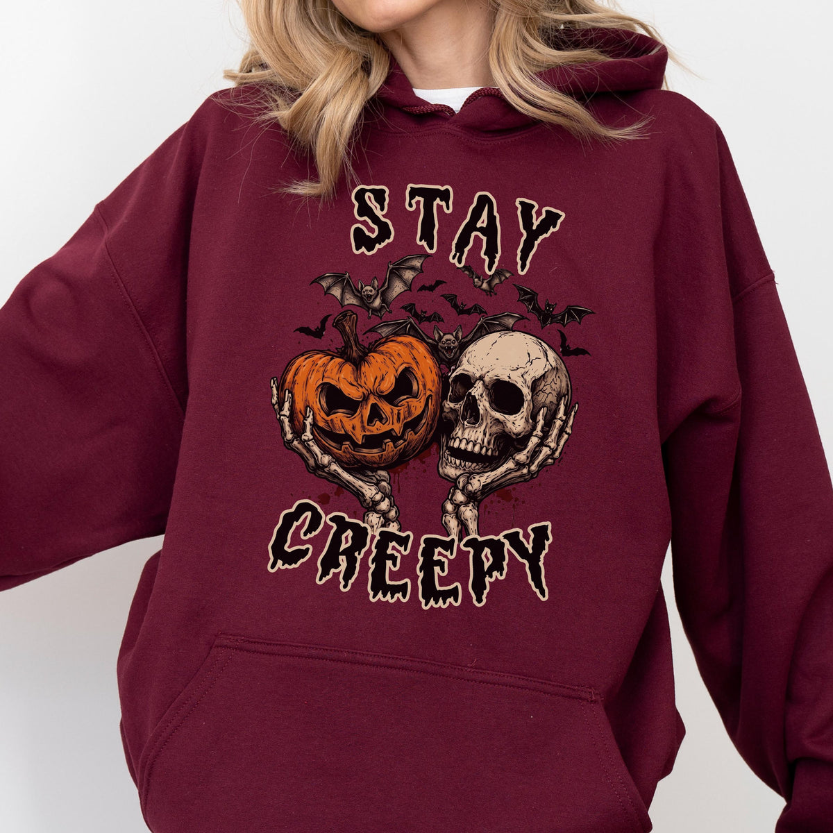 May include: A maroon hooded sweatshirt featuring a graphic print of two skeletal hands holding an orange jack-o'-lantern and a human skull. Above and below the central illustration, the text reads STAY and CREEPY in a dark, dripping font.