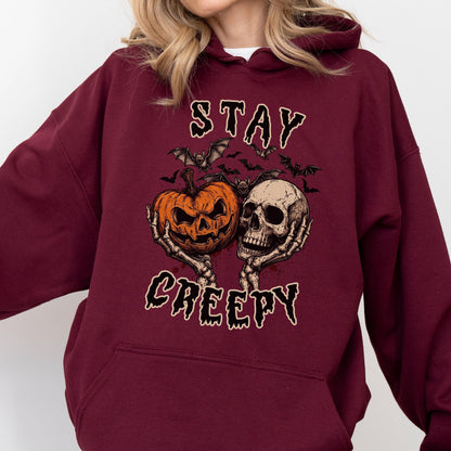 May include: A maroon hooded sweatshirt featuring a graphic print of two skeletal hands holding an orange jack-o'-lantern and a human skull. Above and below the central illustration, the text reads STAY and CREEPY in a dark, dripping font.