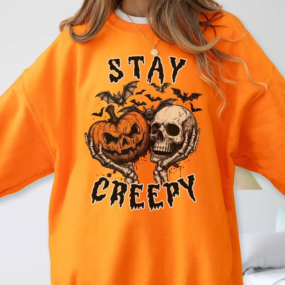 May include: A bright orange crewneck sweatshirt featuring a graphic print of a jack-o'-lantern and a human skull held by skeletal hands. Above and below the illustration, the text reads STAY CREEPY in a black, jagged, distressed font.