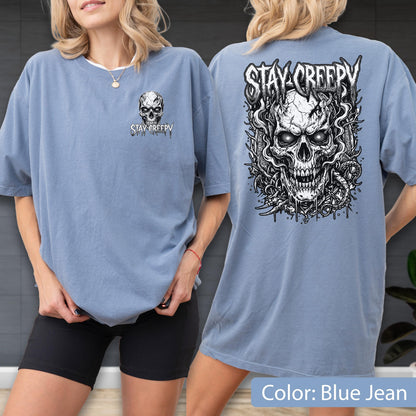 May include: A blue jean colored t-shirt featuring a small skull graphic on the front left chest with the text STAY CREEPY. The back of the shirt displays a large, detailed black and white illustration of a skull surrounded by smoke and vines.