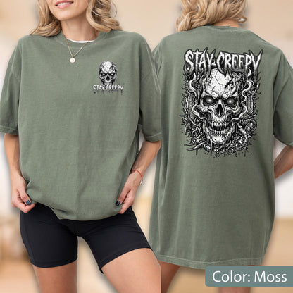 May include: An olive green t-shirt featuring a small skull graphic and the text "STAY CREEPY" on the front left chest. The back of the shirt displays a large, detailed black and white illustration of a skull surrounded by smoke and vines.