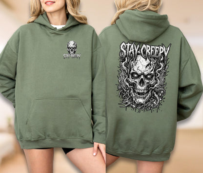 May include: An olive green hooded sweatshirt featuring a small skull graphic and the text STAY CREEPY on the front left chest. The back displays a large, detailed black and white illustration of a skull surrounded by smoke and the text STAY CREEPY.