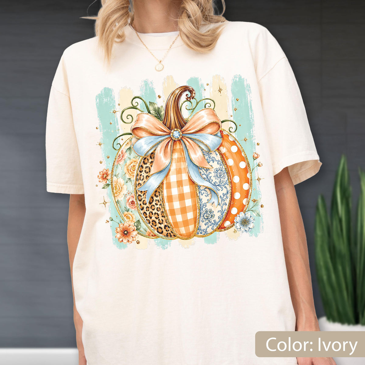 May include: An ivory t-shirt featuring a large graphic of a pumpkin decorated with a light blue bow. The pumpkin design includes sections with leopard print, orange gingham, floral patterns, and white polka dots against a light blue brushstroke background.