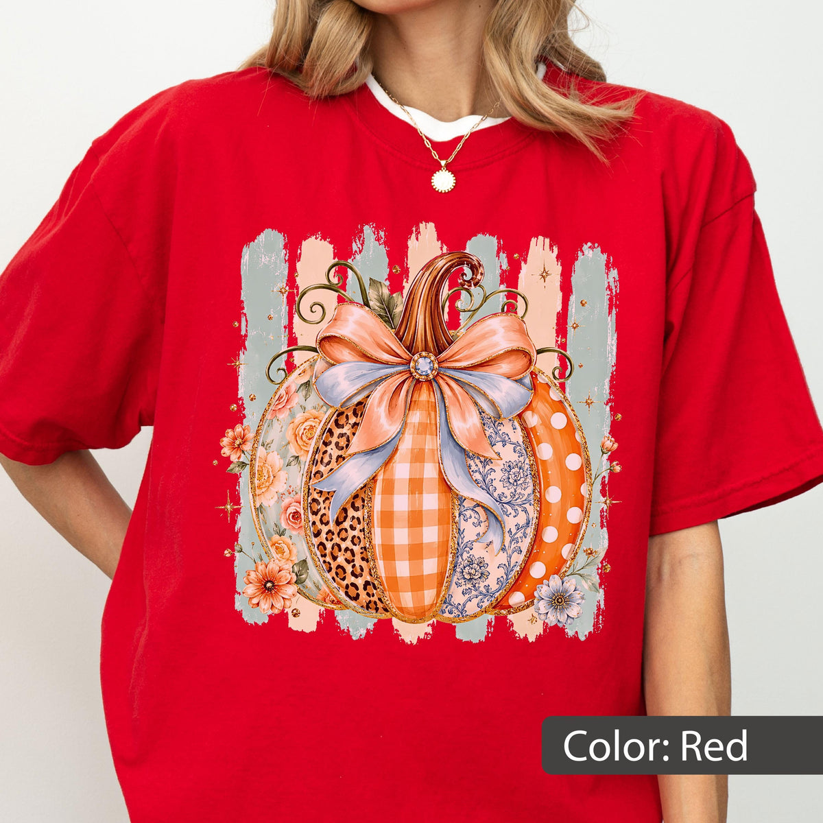 May include: A bright red short-sleeved t-shirt featuring a central graphic of a pumpkin decorated with a large light blue bow. The pumpkin design incorporates various patterns including leopard print, orange gingham, floral motifs, and white polka dots.