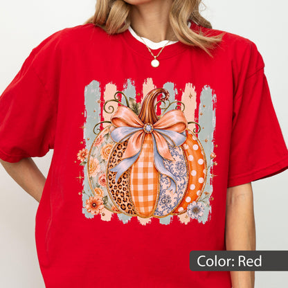May include: A bright red short-sleeved t-shirt featuring a central graphic of a pumpkin decorated with a large light blue bow. The pumpkin design incorporates various patterns including leopard print, orange gingham, floral motifs, and white polka dots.