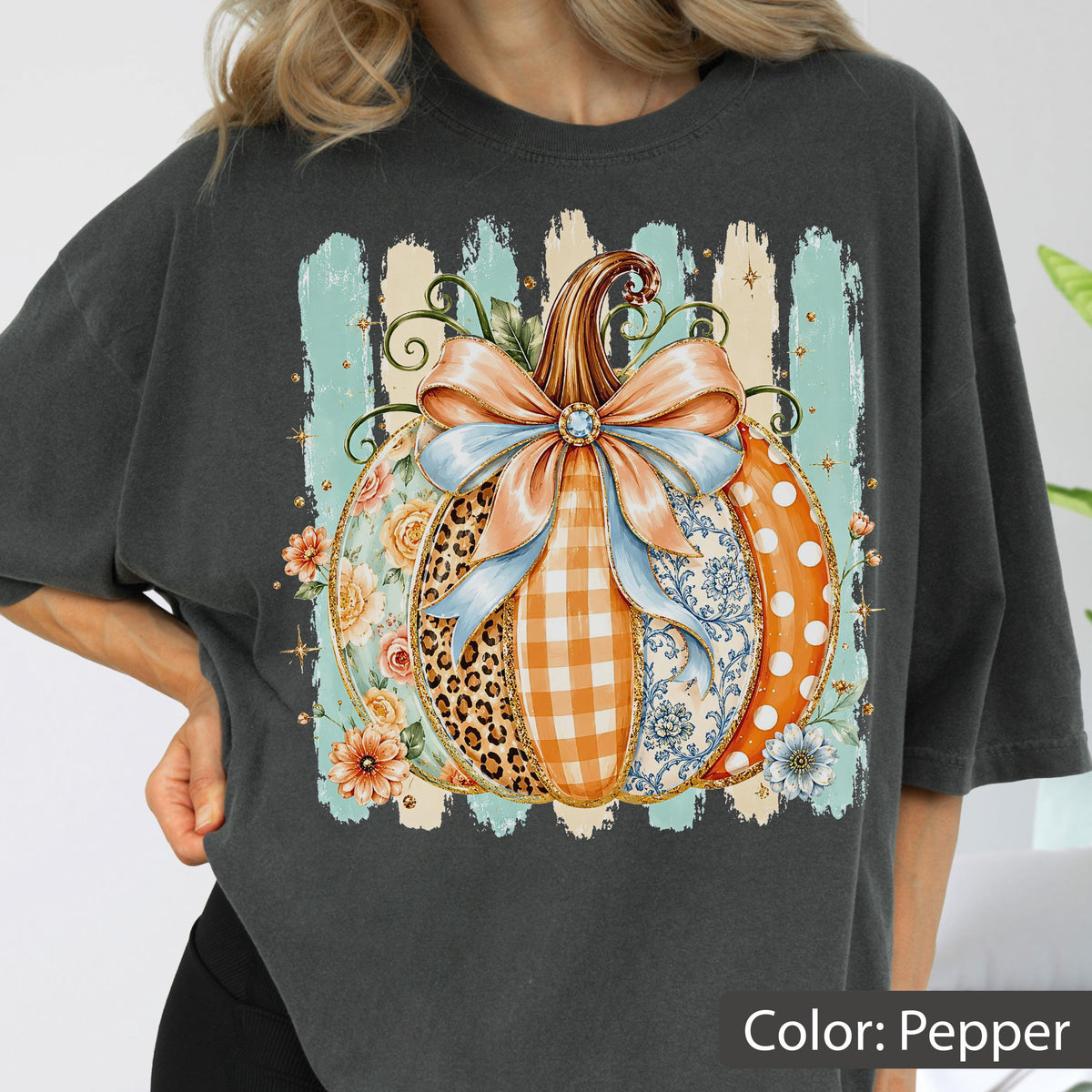 May include: A charcoal gray t-shirt featuring a large graphic of a pumpkin decorated with a light blue bow. The pumpkin segments display various patterns including leopard print, orange gingham, floral motifs, and white polka dots on orange.
