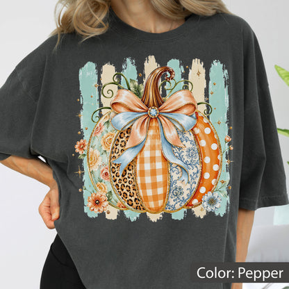 May include: A charcoal gray t-shirt featuring a large graphic of a pumpkin decorated with a light blue bow. The pumpkin segments display various patterns including leopard print, orange gingham, floral motifs, and white polka dots on orange.