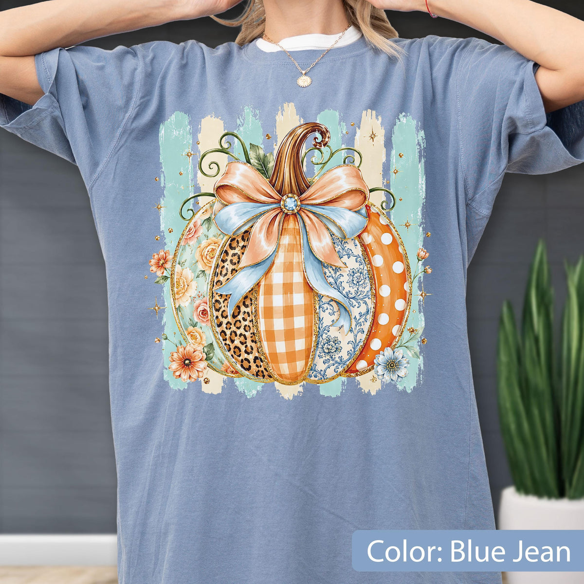 May include: A blue jean colored t-shirt featuring a large graphic of a pumpkin decorated with various patterns including leopard print, orange gingham, and blue floral designs. The pumpkin is topped with a large light blue bow and surrounded by flowers.