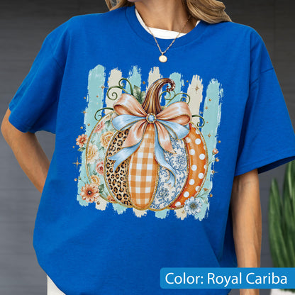 May include: A royal blue t-shirt featuring a central graphic of a pumpkin decorated with various patterns including leopard print, orange gingham, blue floral, and white polka dots, topped with a large light blue bow and surrounded by flowers.