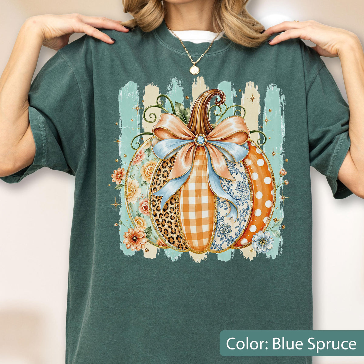 May include: A dark teal t-shirt featuring a large graphic of a pumpkin decorated with a light blue bow. The pumpkin segments display various patterns including leopard print, orange gingham, floral motifs, and white polka dots on an orange background.