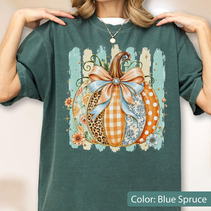 May include: A dark teal t-shirt featuring a large graphic of a pumpkin decorated with a light blue bow. The pumpkin segments display various patterns including leopard print, orange gingham, floral motifs, and white polka dots on an orange background.