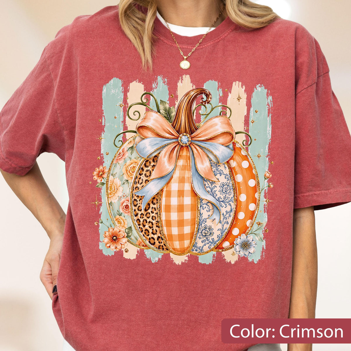 May include: A crimson short-sleeve t-shirt featuring a central graphic of a pumpkin decorated with a large light blue bow. The pumpkin design incorporates various patterns including leopard print, orange gingham, floral motifs, and white polka dots.