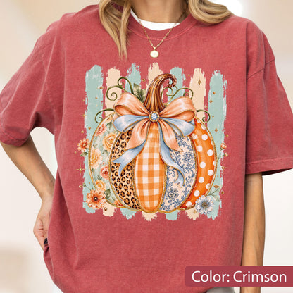 May include: A crimson short-sleeve t-shirt featuring a central graphic of a pumpkin decorated with a large light blue bow. The pumpkin design incorporates various patterns including leopard print, orange gingham, floral motifs, and white polka dots.
