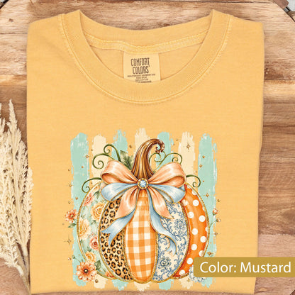 May include: A mustard yellow t-shirt featuring a graphic of a pumpkin decorated with a large light blue bow. The pumpkin design includes sections with leopard print, orange gingham, floral patterns, and white polka dots on a light blue brushstroke background.