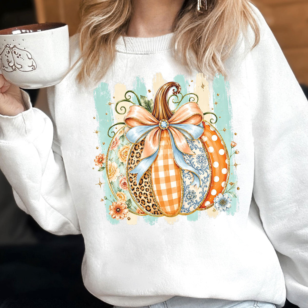 May include: A white crewneck sweatshirt featuring a central graphic of a pumpkin decorated with various patterns including leopard print, orange gingham, and floral motifs. The pumpkin is adorned with a large light blue bow and sits against a light teal striped background.