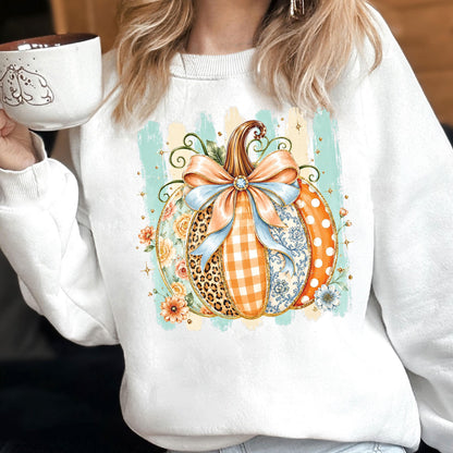May include: A white crewneck sweatshirt featuring a central graphic of a pumpkin decorated with various patterns including leopard print, orange gingham, and floral motifs. The pumpkin is adorned with a large light blue bow and sits against a light teal striped background.